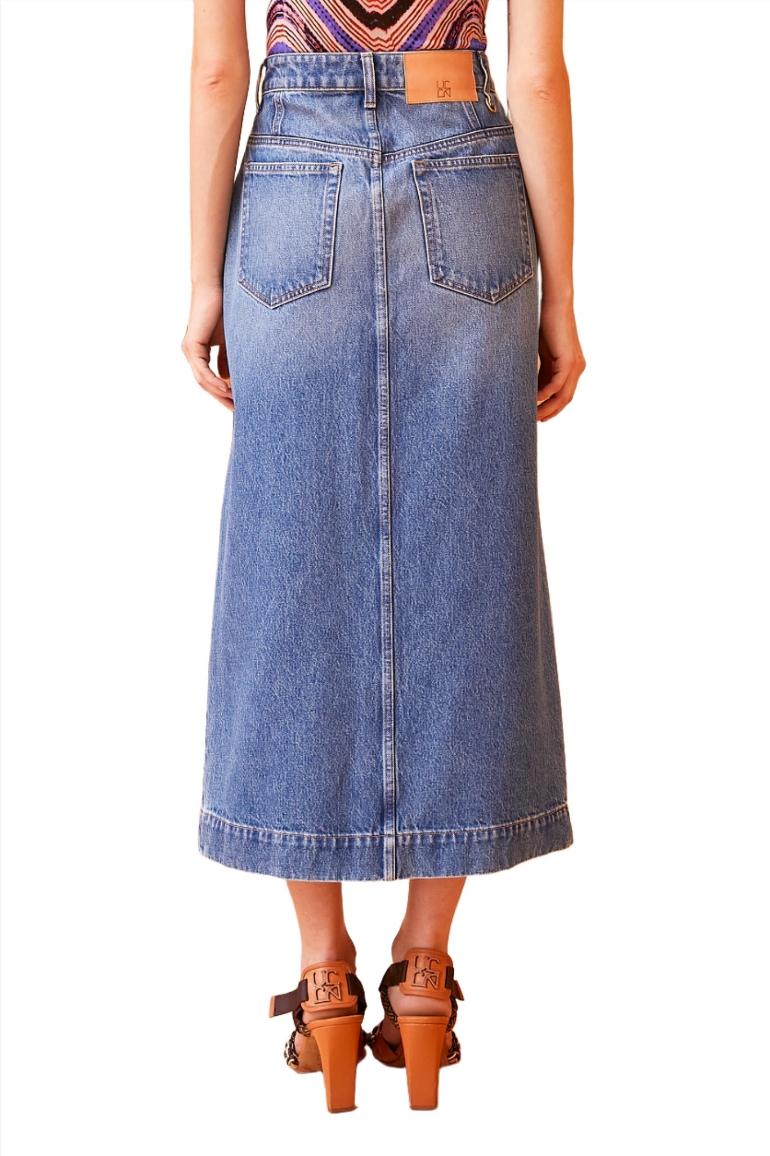 A person stands with their back facing the camera, wearing an Ulla Johnson Bea Skirt featuring a front split made from non-stretch denim, paired with brown heels.