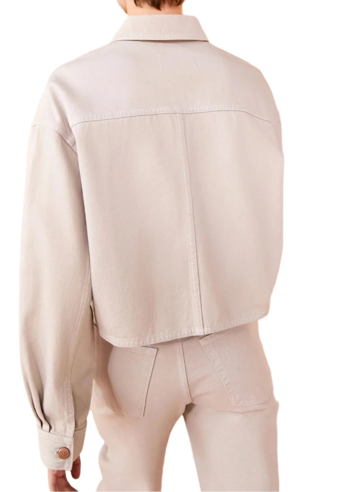 A person is shown from the back wearing Ulla Johnson The Bobbi Denim Jacket in light beige, paired with matching high-waisted pants. The rigid denim jacket features a wide back yoke and buttoned cuffs, exuding a crisp, structured elegance.