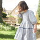 A young girl in a Pink Chicken Girls' Maribelle Dress with a blue climbing floral print on soft cotton poplin stands outside on grass, backed by stone buildings and trees.