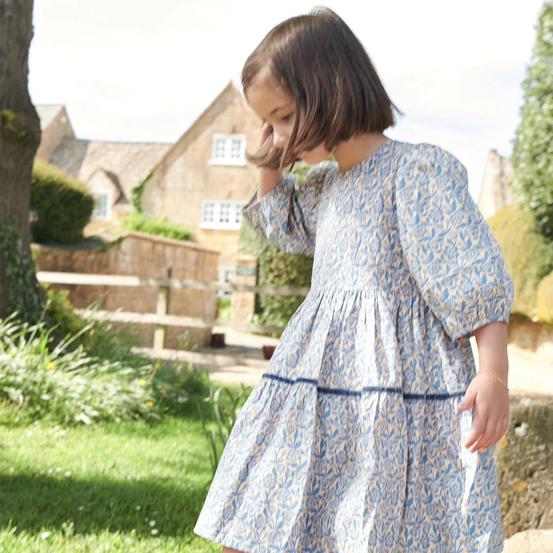 A young girl in a Pink Chicken Girls' Maribelle Dress with a blue climbing floral print on soft cotton poplin stands outside on grass, backed by stone buildings and trees.