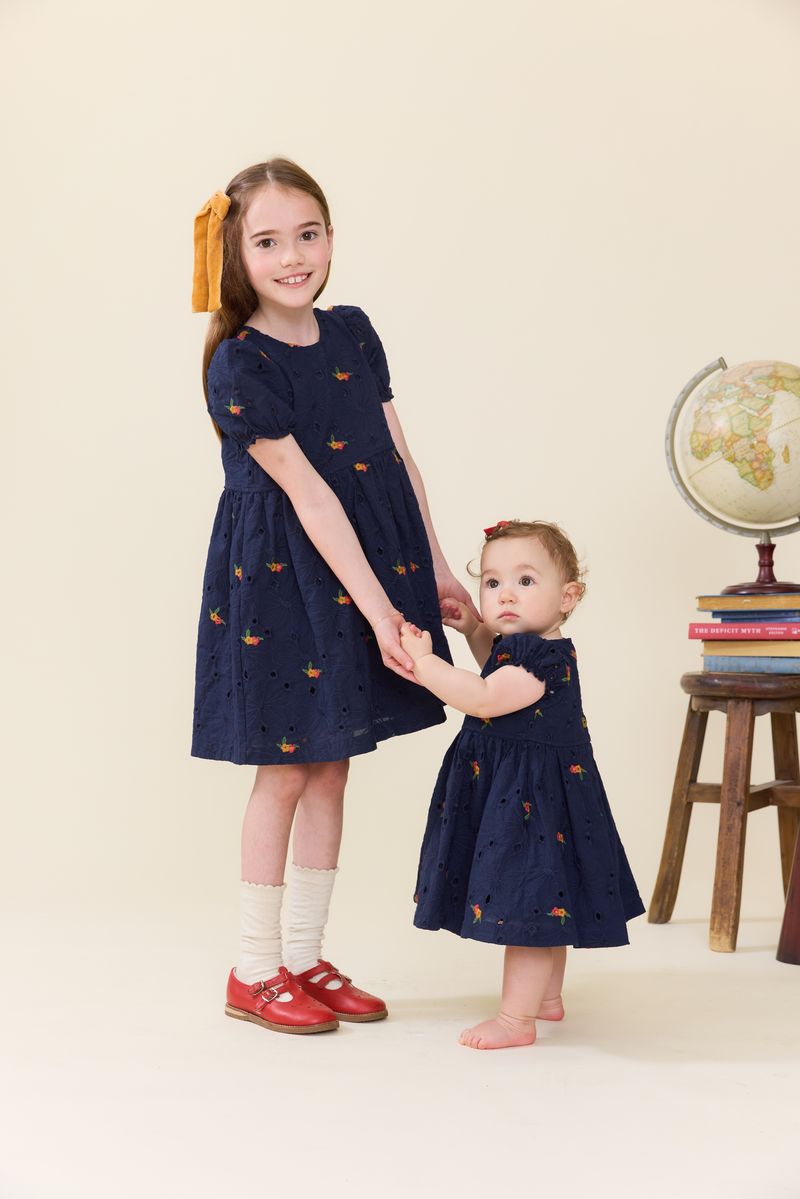 Two young girls—one older with a yellow hair bow, the other a toddler—hold hands, both dressed in matching navy blue Baby Girls' Eliza Eyelet Embroidery Dress Sets by Pink Chicken, which feature delicate navy floral embroidery. A globe and stacked books rest on a stool nearby.