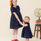 Wearing matching Pink Chicken Girls' Eliza Eyelet Embroidery Dresses, two young girls hold hands. The older girl grins warmly, while the younger gazes forward. A globe and a stack of books rest on a stool nearby, evoking the timeless charm of a classic fall dress scene.