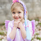 A young girl smiles outdoors with her hands under her chin, wearing the Great Pretenders Unicorn Princess Hair Braid and a pastel unicorn costume with wings.
