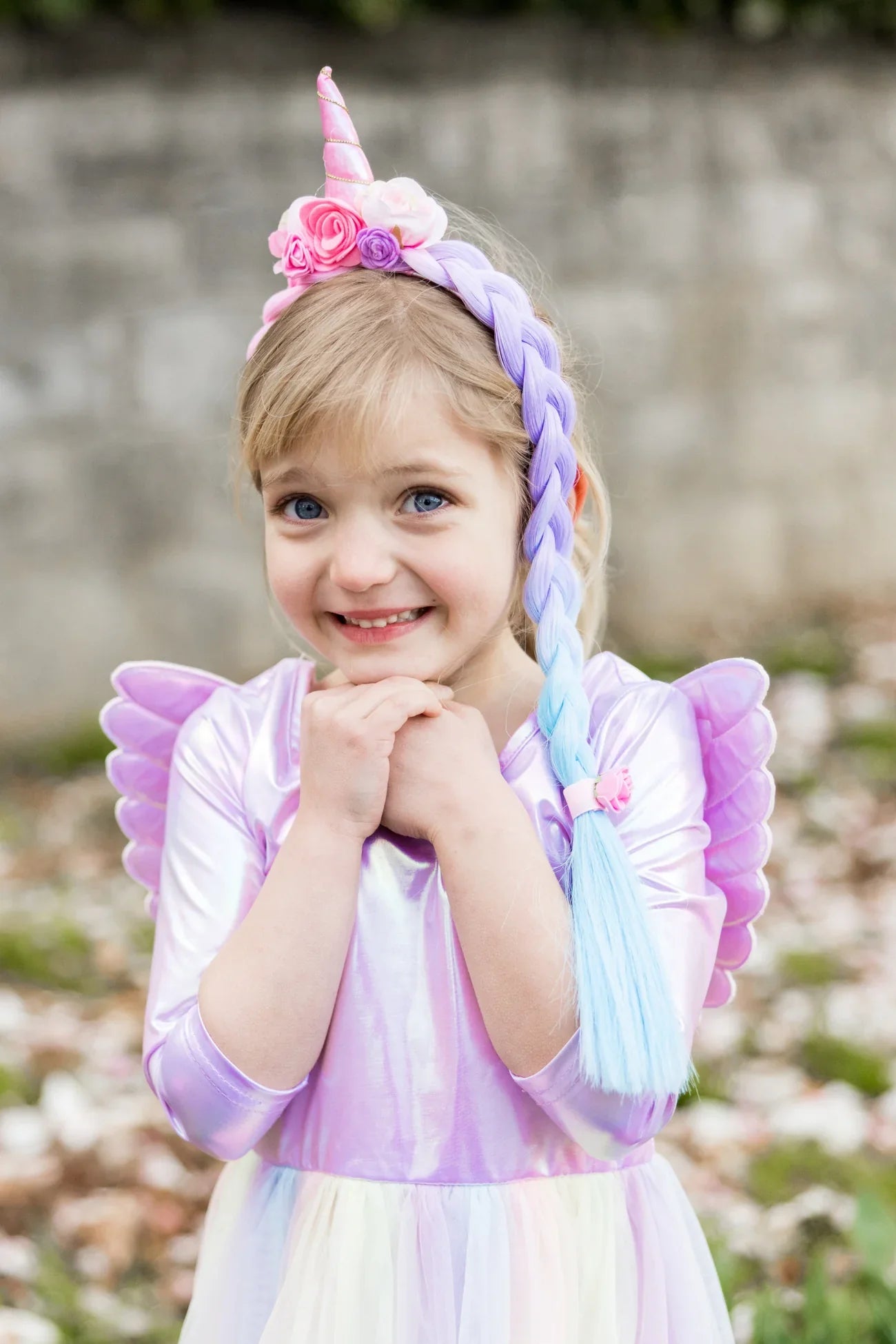 A young girl smiles outdoors with her hands under her chin, wearing the Great Pretenders Unicorn Princess Hair Braid and a pastel unicorn costume with wings.