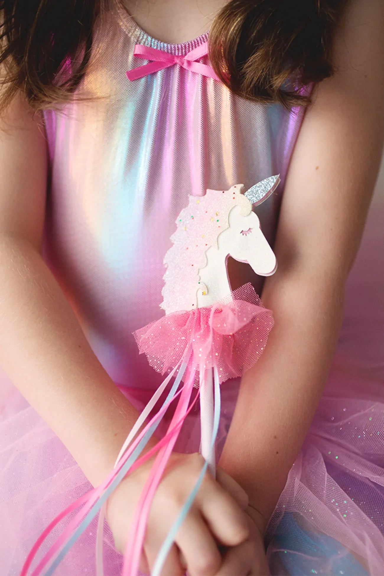 A girl in a shiny pink dress holds the Great P Fanciful Unicorn Wand by Great Pretenders, adorned with ribbons and a tutu, ready for fairy-tale fun.