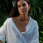 A woman with long dark hair, dressed in a white button-up shirt, wears the Anabel Aram Bamboo Necklace and sits before lush green tropical plants.
