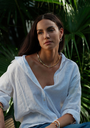 A woman with long dark hair, dressed in a white button-up shirt, wears the Anabel Aram Bamboo Necklace and sits before lush green tropical plants.