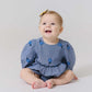 A baby girl sits on the floor, smiling in the Pink Chicken Baby Girls' Rowan Bubble—featuring puffed sleeves, blue blossom embroidery, and a yellow bow—designed for easy diaper changes.