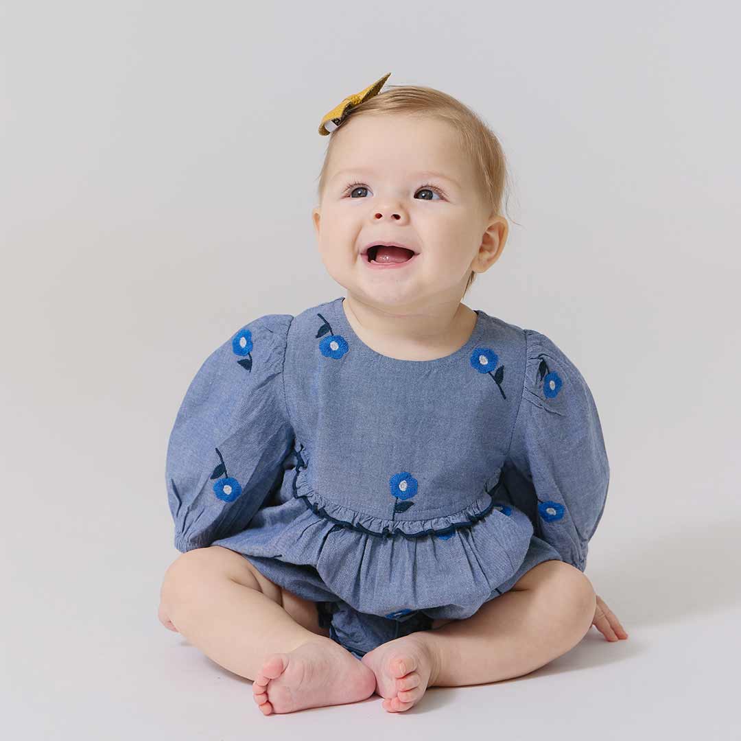 A baby girl sits on the floor, smiling in the Pink Chicken Baby Girls' Rowan Bubble—featuring puffed sleeves, blue blossom embroidery, and a yellow bow—designed for easy diaper changes.