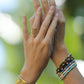Two hands adorned with gold jewelry, including the Anabel Aram Bamboo Wide Cuff and a textured ring, plus several colorful beaded bracelets, are held up against a blurred green background.