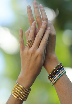 Two hands adorned with gold jewelry, including the Anabel Aram Bamboo Wide Cuff and a textured ring, plus several colorful beaded bracelets, are held up against a blurred green background.