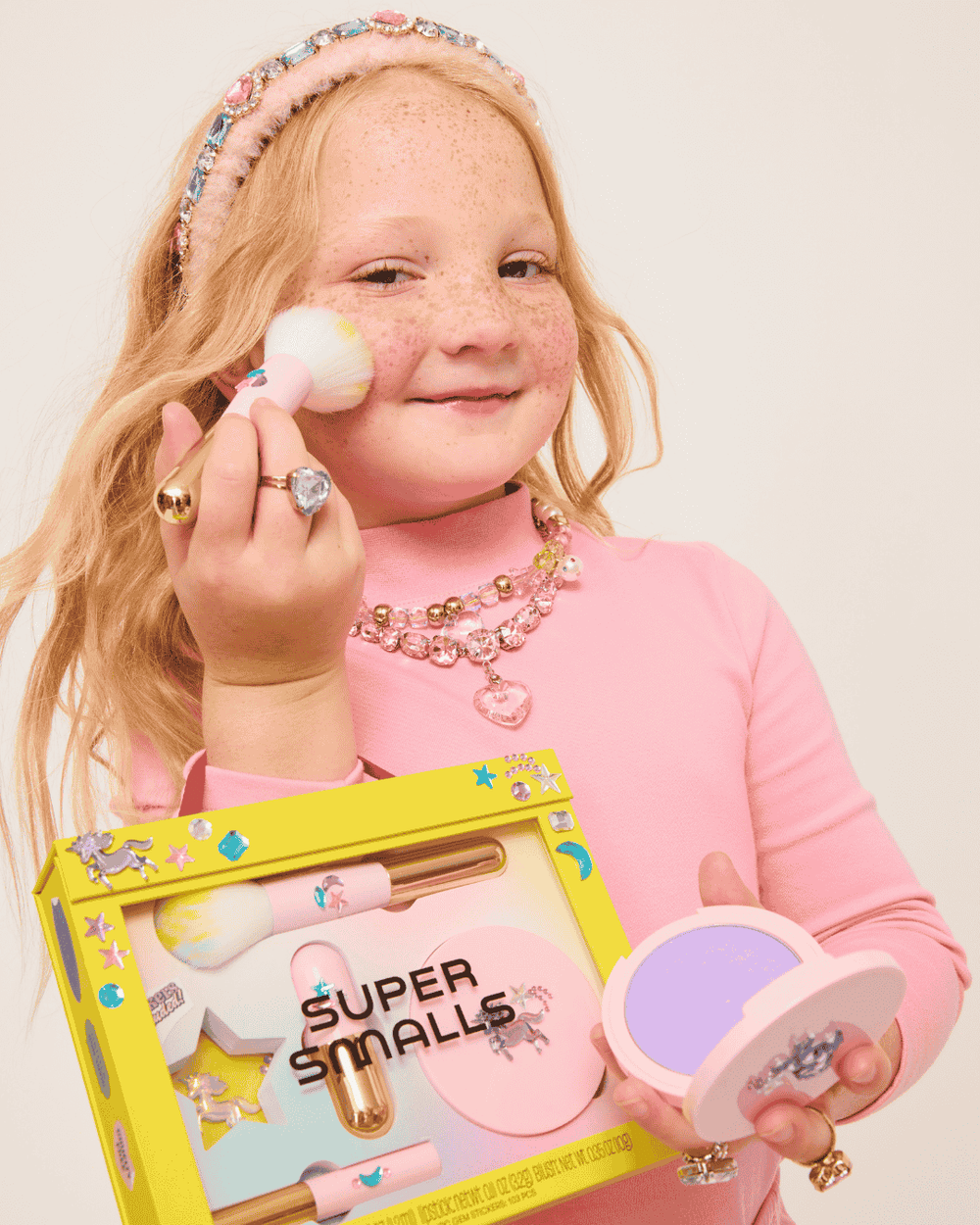 A smiling young girl with freckles applies the Super Smalls Unicorn Makeup Kit using a brush, dressed in pink with playful jewelry and a headband—ideal for imaginative play.