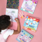 A child colors a Floss and Rock Magic Colouring Fun - Dinosaur book with colorful markers on a pink surface, surrounded by three spiral-bound books, discovering the surprise of invisible ink magic colouring-in.
