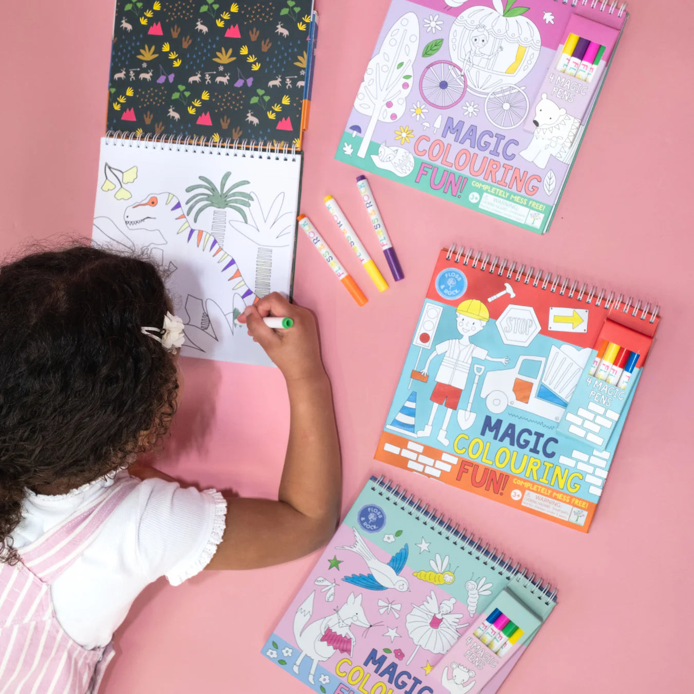 A child colors a Floss and Rock Magic Colouring Fun - Dinosaur book with colorful markers on a pink surface, surrounded by three spiral-bound books, discovering the surprise of invisible ink magic colouring-in.