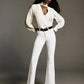 A woman poses in a studio, styled in FRAME's The Icon full-length flared jeans—a white super-stretch denim that channels the iconic '70s silhouette—paired with a cream blouse, black belt, and white heeled sandals.
