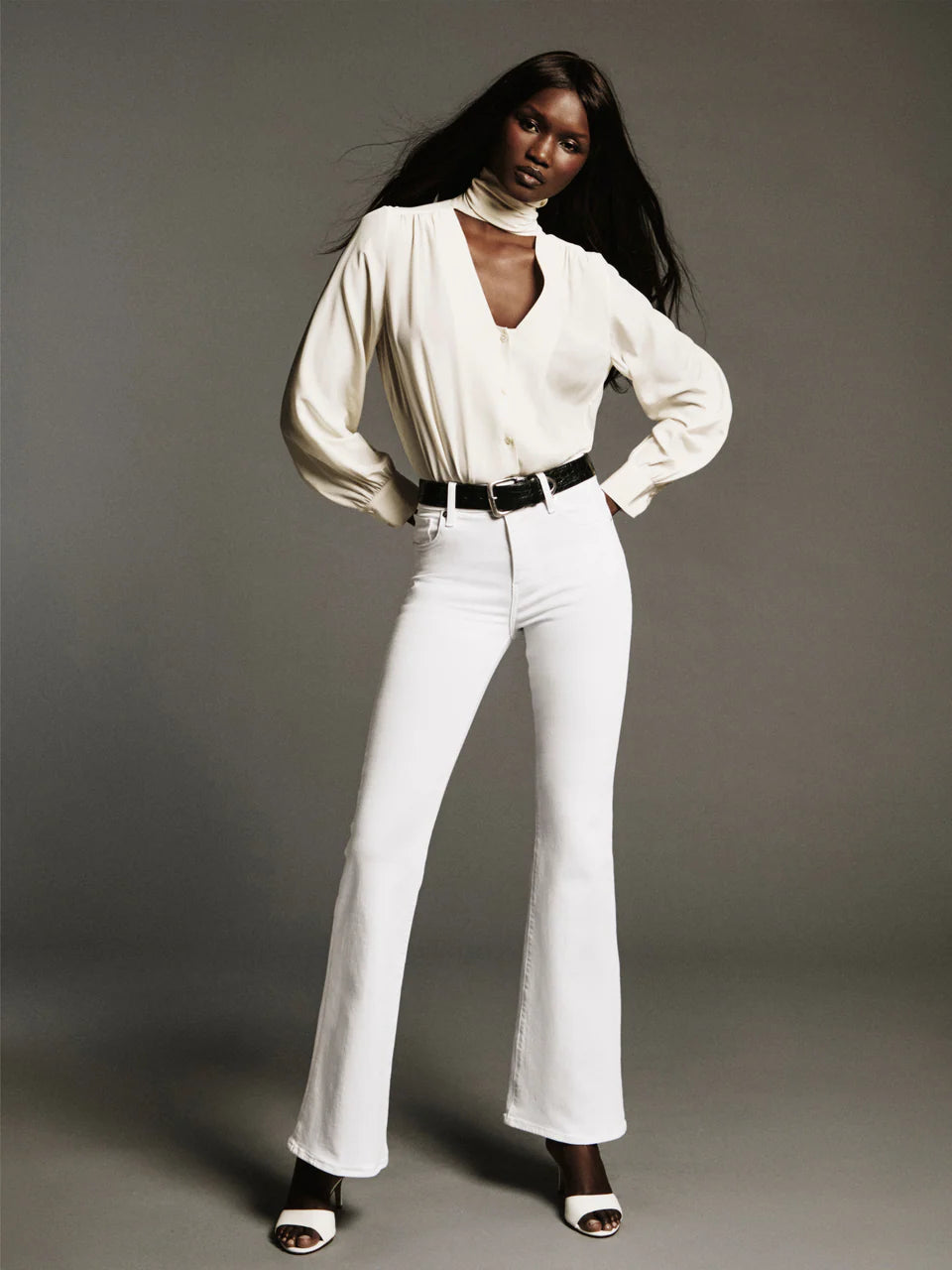 A woman poses in a studio, styled in FRAME's The Icon full-length flared jeans—a white super-stretch denim that channels the iconic '70s silhouette—paired with a cream blouse, black belt, and white heeled sandals.