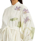 A woman wears the Erdem Long Sleeve Midi Dress With Tie Waist Detail, featuring a white linen design with sheer sleeves and purple and green botanical illustrations.
