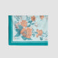 The Trailing Poppy Pareo by Erdem is a light blue scarf with orange and green poppy floral print, a teal border, and "ERDEM" in one corner, crafted from a soft silk-modal blend.