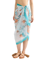 A person wearing the Trailing Poppy Pareo by Erdem, a light blue sarong with a poppy print, crafted from a soft silk-modal blend and tied at the waist, paired with brown tasseled sandals.