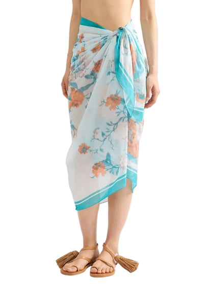 A person wearing the Trailing Poppy Pareo by Erdem, a light blue sarong with a poppy print, crafted from a soft silk-modal blend and tied at the waist, paired with brown tasseled sandals.