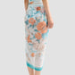 A woman wears the Erdem Trailing Poppy Pareo—a blue floral-patterned sheer sarong skirt—paired with a matching bikini top and tassel-trimmed flat sandals, standing against a plain background.