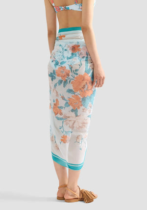 A woman wears the Erdem Trailing Poppy Pareo—a blue floral-patterned sheer sarong skirt—paired with a matching bikini top and tassel-trimmed flat sandals, standing against a plain background.