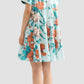 A woman faces away, wearing the Erdem Trailing Poppy Short Shirt Dress—a loose, short-sleeved light blue cotton sateen mini dress with orange and white poppy print—paired with brown tassel sandals.