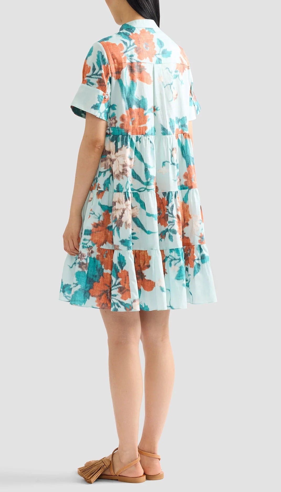 A woman faces away, wearing the Erdem Trailing Poppy Short Shirt Dress—a loose, short-sleeved light blue cotton sateen mini dress with orange and white poppy print—paired with brown tassel sandals.
