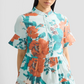 A woman wears the Erdem Trailing Poppy Short Shirt Dress, a light blue cotton sateen mini dress with orange and teal poppy print, mandarin collar, short ruffled sleeves, and a button-down front.