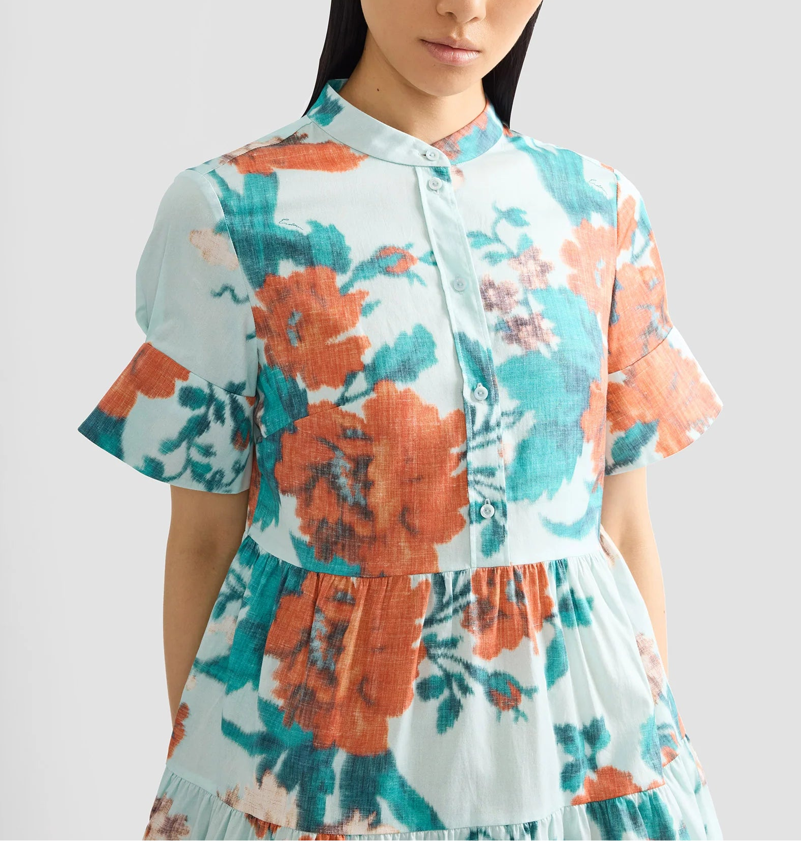 A woman wears the Erdem Trailing Poppy Short Shirt Dress, a light blue cotton sateen mini dress with orange and teal poppy print, mandarin collar, short ruffled sleeves, and a button-down front.
