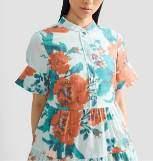 A woman wears the Erdem Trailing Poppy Short Shirt Dress, a light blue cotton sateen mini dress with orange and teal poppy print, mandarin collar, short ruffled sleeves, and a button-down front.