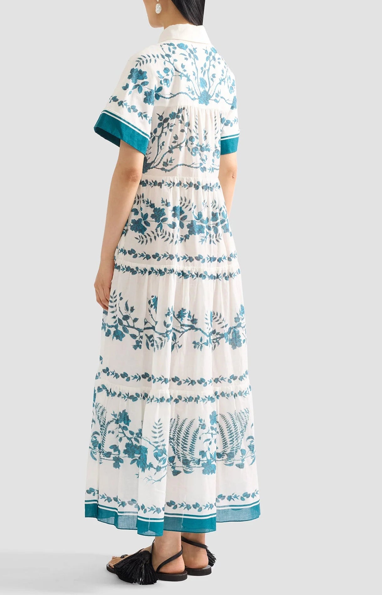 A woman stands facing away, wearing the Erdem Wild Fern Botanical Shirt Dress featuring a teal fern design and short sleeves, paired with flat black sandals.