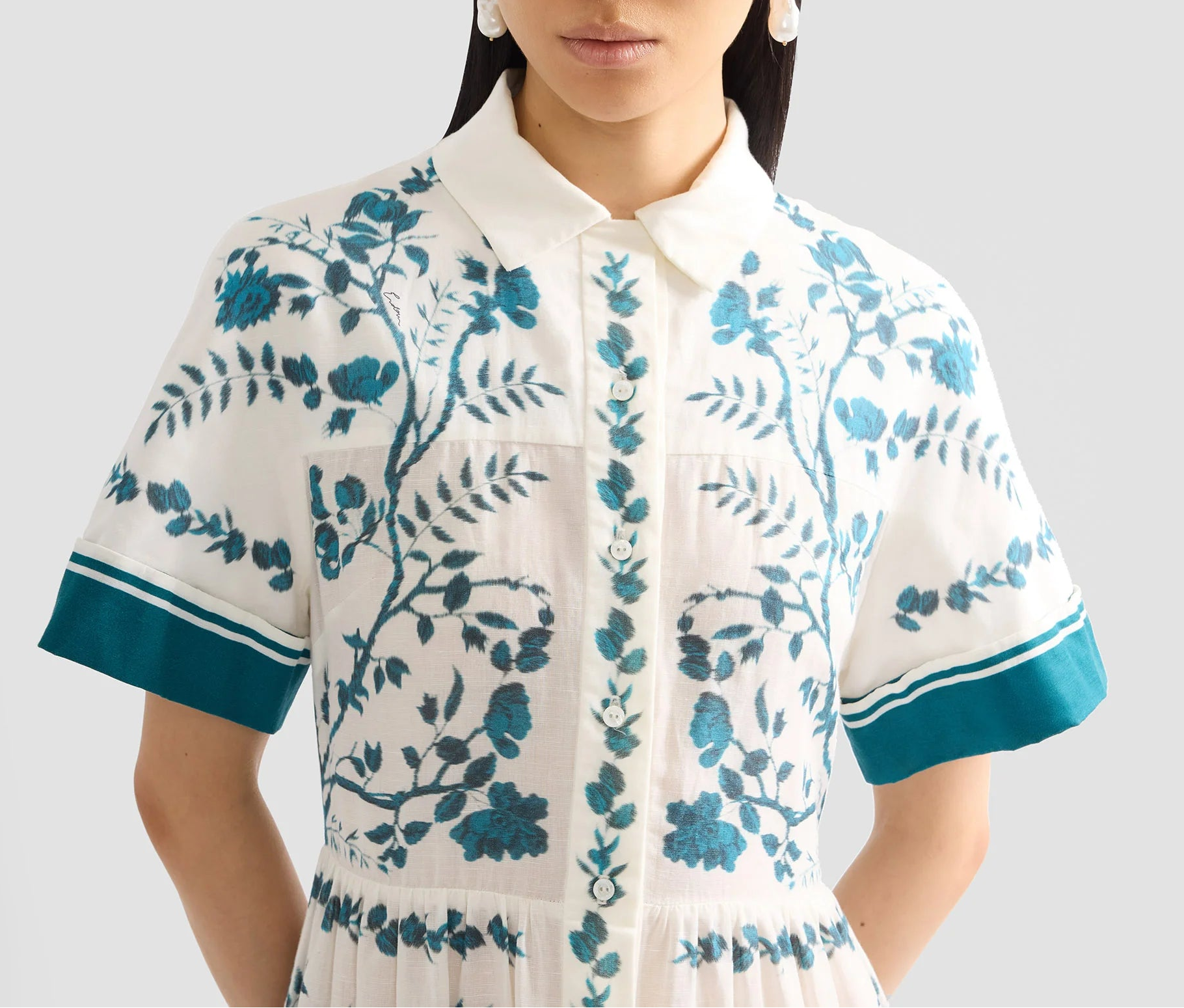 A woman wears the Erdem Wild Fern Botanical Shirt Dress, an ankle-length white cotton linen-blend dress with teal floral embroidery and matching teal trim on the short sleeves.