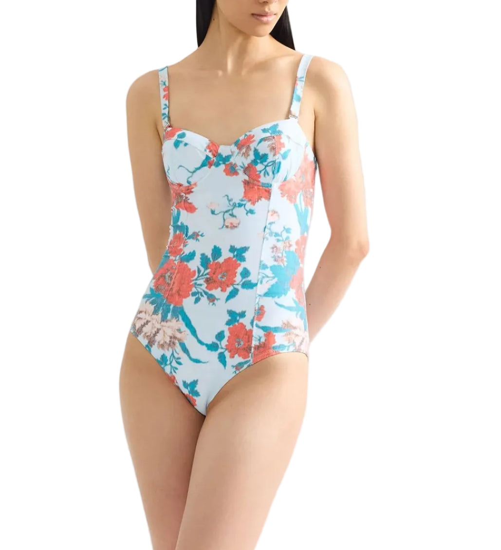 A woman wears the Erdem Trailing Poppy One Piece, a light blue swimsuit with red and pink floral patterns, moulded cups, thin shoulder straps, and a sweetheart neckline.