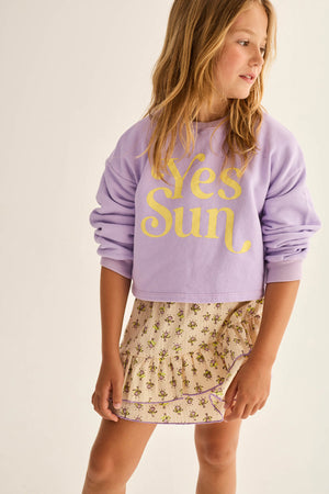 A young girl wears the Cozmo Girls' Vega Sweatshirt with a crew neckline and "Yes Sun" in yellow, paired with a beige floral skirt, as she stands slightly turned to the side.