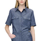 The individual wears the Vesper Top by Callas Milano, a short-sleeve denim jumpsuit in Indigo with buttoned pockets, standing against a plain white background. The outfit radiates timeless retro appeal.