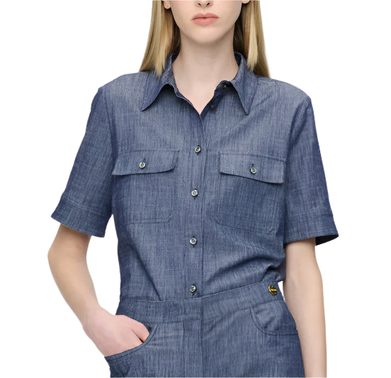 The individual wears the Vesper Top by Callas Milano, a short-sleeve denim jumpsuit in Indigo with buttoned pockets, standing against a plain white background. The outfit radiates timeless retro appeal.
