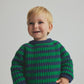 A toddler with blond hair, wearing the Cozmo Baby Boys’ Vigo Knit Striped Sweater in green and blue along with matching blue pants, stands against a plain light background, looking slightly to the side and smiling.