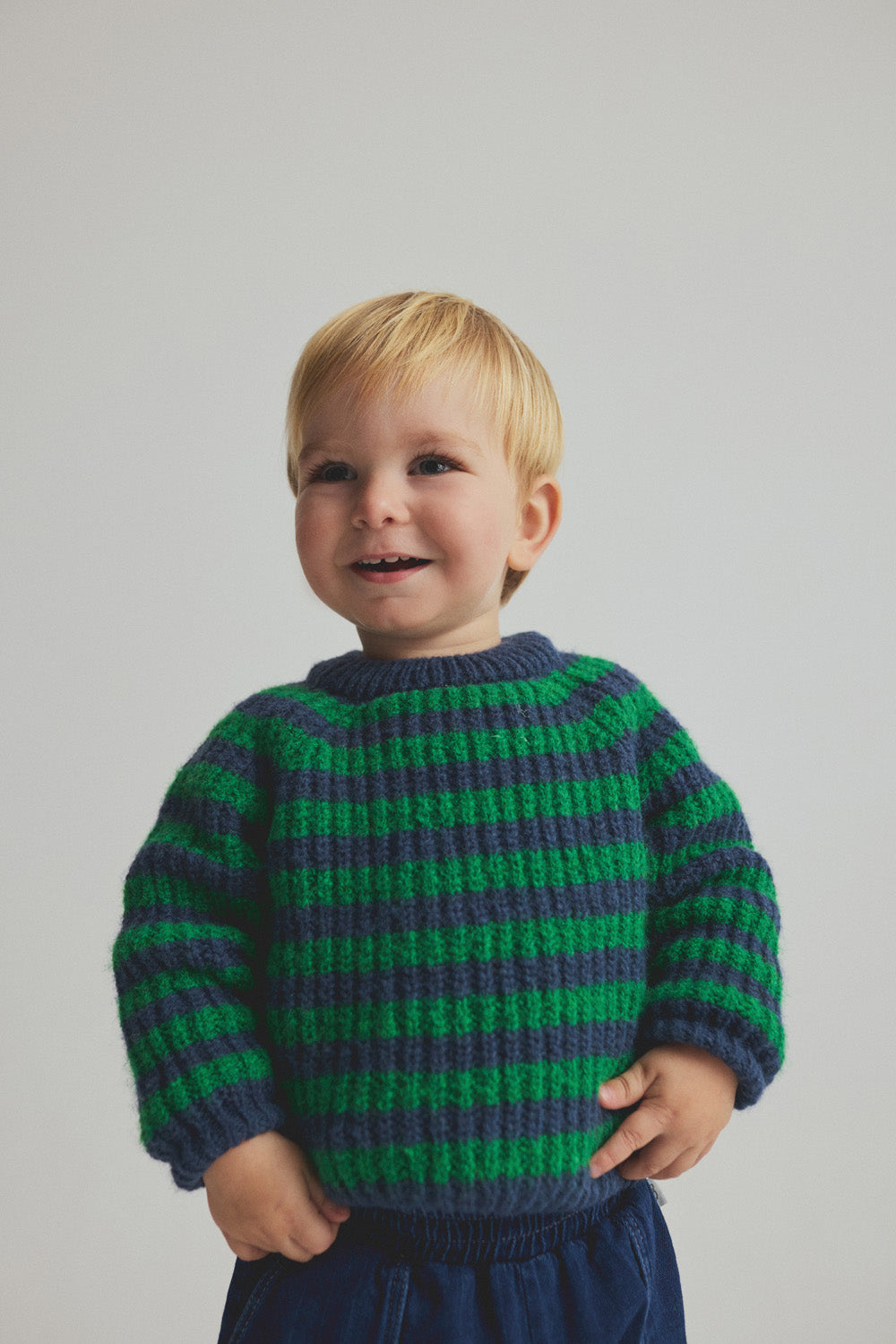 A toddler with blond hair, wearing the Cozmo Baby Boys’ Vigo Knit Striped Sweater in green and blue along with matching blue pants, stands against a plain light background, looking slightly to the side and smiling.