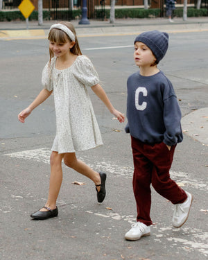 Two children cross a street; the girl wears Rylee & Cru’s Girls' Sabrina Mini Dress with a headband, while the boy wears a beanie, dark sweatshirt with a "C," and maroon pants.