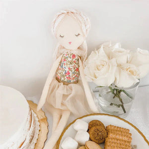 The MON AMI Vanilla Scented Doll from Mon Ami sits beside a white cake, a vase of white roses, and a plate of assorted cookies and marshmallows. Vanilla cupcakes add to the delightful setting, infusing the atmosphere with their sweet aroma.