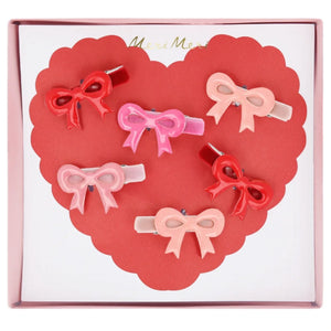 Six Meri Meri Valentine's Bow Hair Clips, in velvet shades of red, pink, and peach, are beautifully arranged on a heart-shaped backing in a box, perfect for adding charm this Valentine's Day.