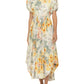 A woman wearing the Aire Maxi Dress by Agua By Aguabendita, a light floral A-line dress with puff sleeves and an asymmetrical hem, paired with silver sandals.