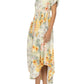 A person wearing the Aire Maxi Dress by Agua By Aguabendita, featuring a floral pattern, asymmetrical hemline, and hand-embroidered details, paired with silver sandals against a plain white background.