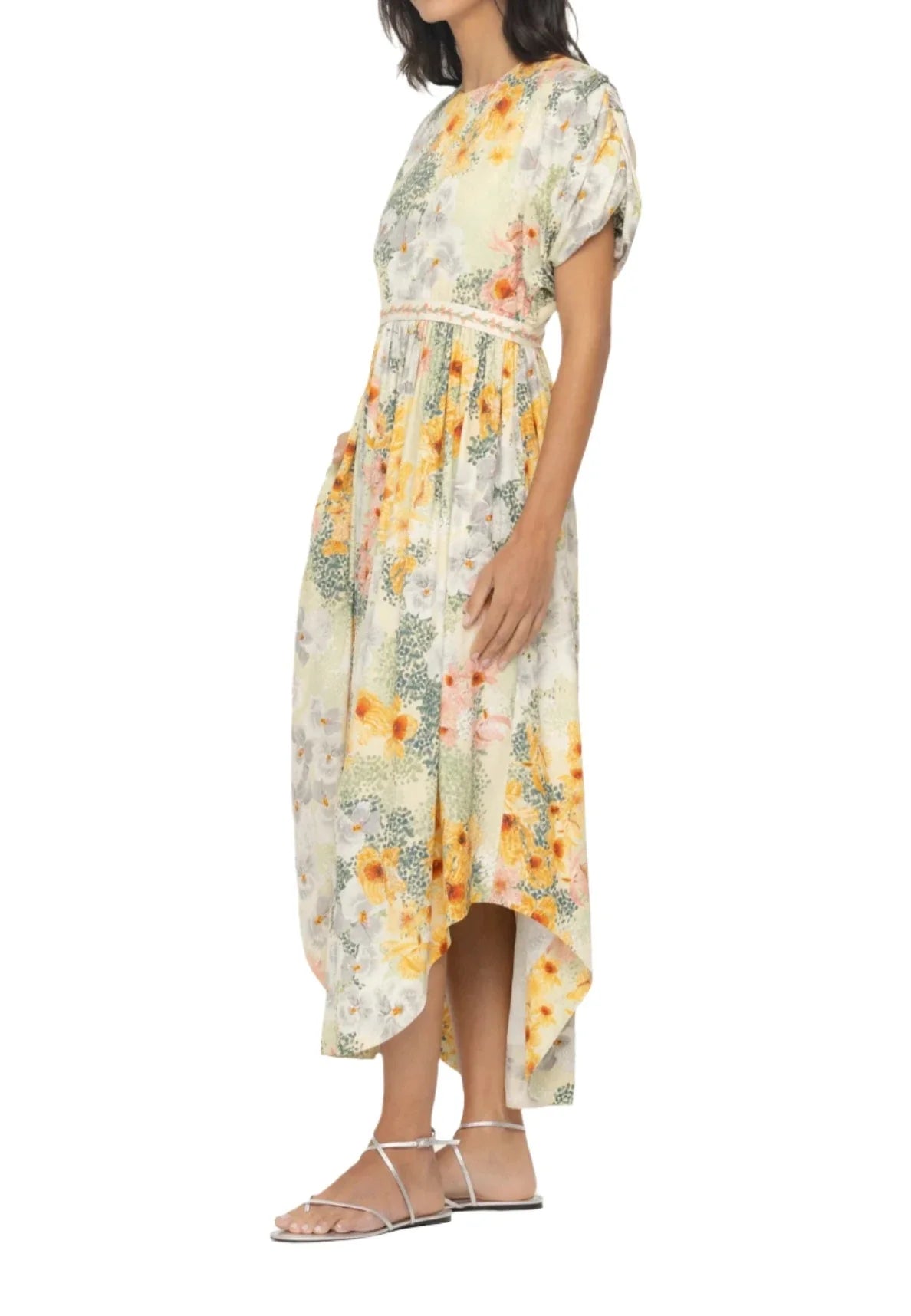 A person wearing an Agua By Aguabendita Aire Maxi Dress with a floral pattern and high-low hem stands with hands in pockets. The A-line silhouette dress features short sleeves, a belted waist, and handmade embroidery accents. White strappy sandals complete the look.