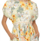 A woman showcases the Aire Maxi Dress by Agua By Aguabendita, featuring an A-line silhouette and puffed short sleeves. The floral design includes watercolor-like shades of yellow, orange, and white. With hands in pockets, this dress exudes effortless charm from shoulders to knees.