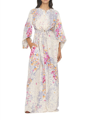 A woman is wearing an Agua By Aguabendita Antonieta Maxi Kaftan made of soft viscose, featuring a floral placement print in pink, purple, and orange tones on a white background. The dress has wide sleeves and a drawstring waist.