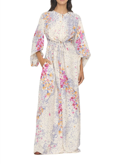 A woman is wearing an Agua By Aguabendita Antonieta Maxi Kaftan made of soft viscose, featuring a floral placement print in pink, purple, and orange tones on a white background. The dress has wide sleeves and a drawstring waist.