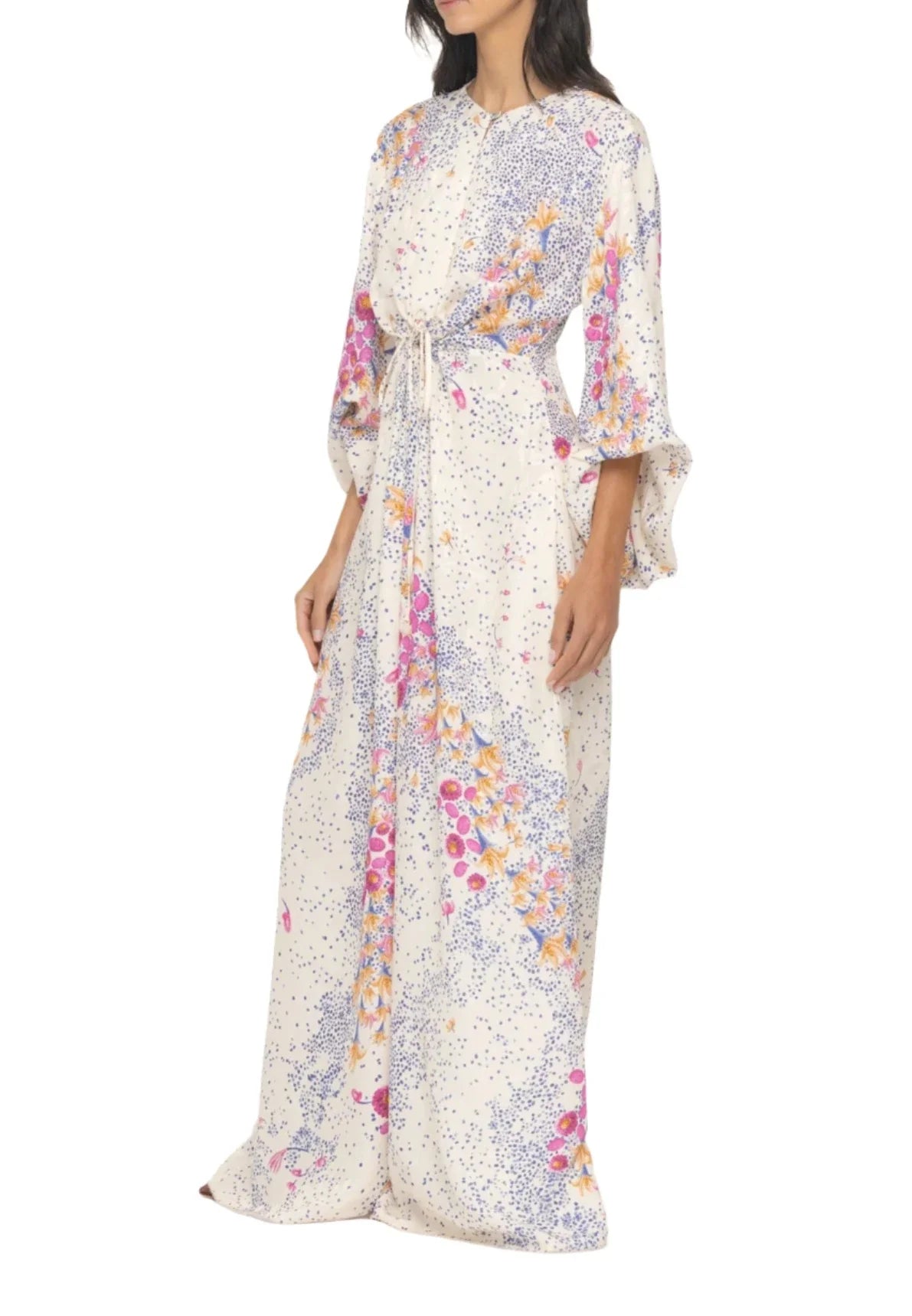 A woman wearing a long, white, floral dress with a colorful placement print and three-quarter sleeves stands with one hand by her side and the other on her hip. The vibrant viscose fabric adds an elegant touch to the Agua By Aguabendita Antonieta Maxi Kaftan style.