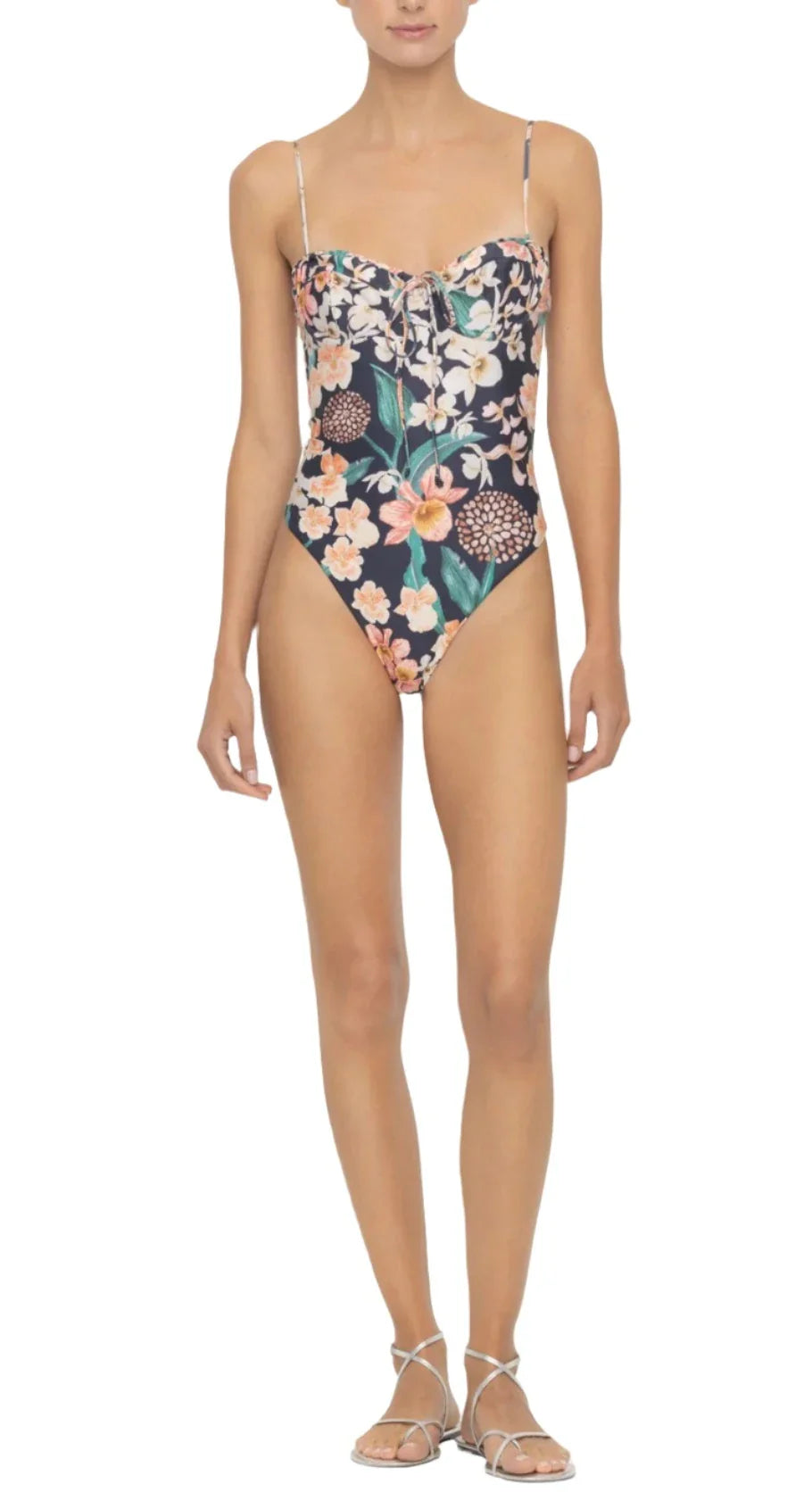 Person wearing the Agua By Aguabendita Ebano One Piece, a floral print swimsuit made from recycled polyester with adjustable straps and a lace-up front, paired with white strappy sandals.